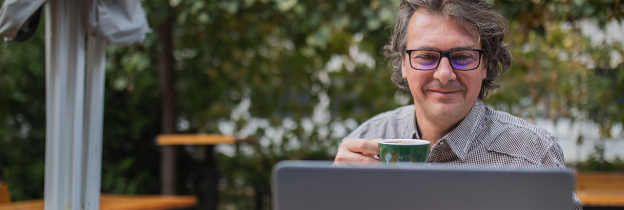 Image d’un homme souriant portant des lunettes, une tasse dans la main droite et consultant son ordinateur portable